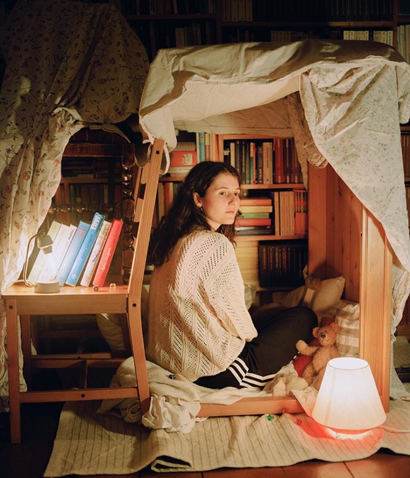 Eine junge Frau sitzt in einer selbstgebauten Höhle aus Stühlen for einem Bücherregal.