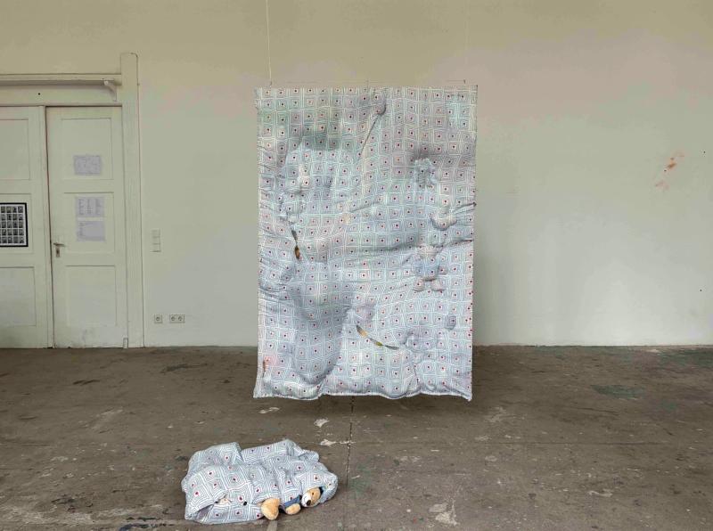 Zu sehen ist ein Werk der Künstlerin Rosa Witt mit den Maßen 70x120x40. Gearbeitet wurde mit Bettwäsche und darin eingenähten Kuscheltieren. Das Werk erinnert an ein aufrecht stehendes Bett. 
