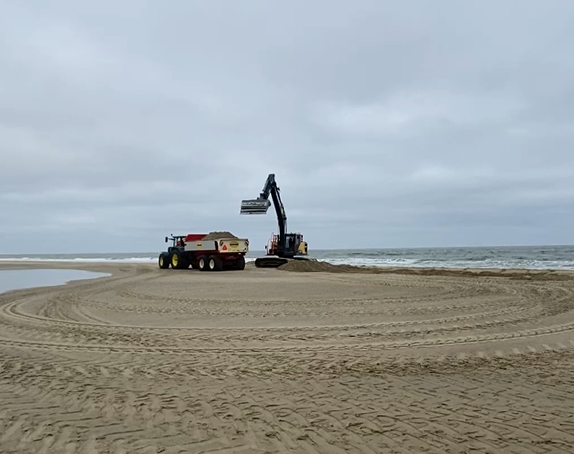 The image shows an excavator on a sandy beach loading sand into a dump truck. The machines are positioned close to the shoreline. Tire marks cover the sand.