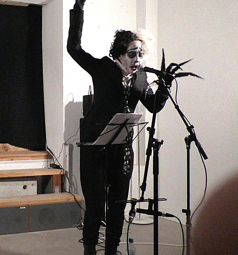 Performerin am Mikrofon, in Kostüm und mit expressivem Make-up, während der Performance „The Burrow Project“.