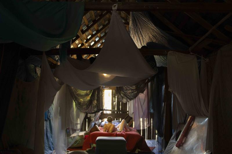 A nude figure lays in a bed. They are surrounded by a dilapedated building. Curtains are hung from the walls and ceiling. 