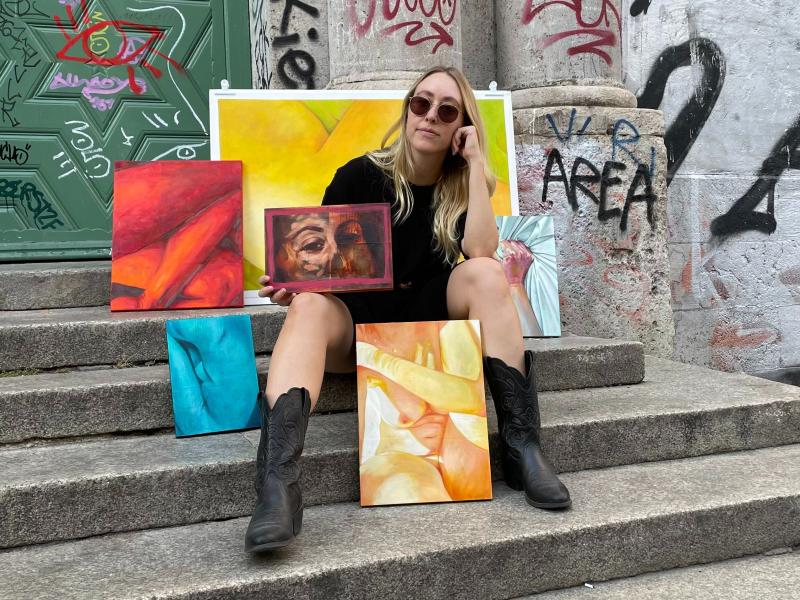 A photograph of the artist Erika Clugston sitting on steps in front of a graffitied building, surrounded by some of her oil paintings.