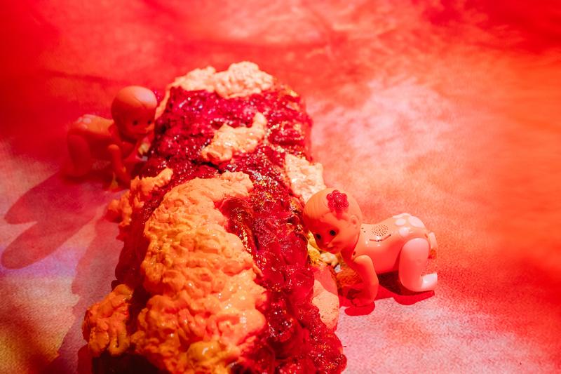Toy baby figures crawl around a red, fleshy sculptural object on a floor with a printed nipple.