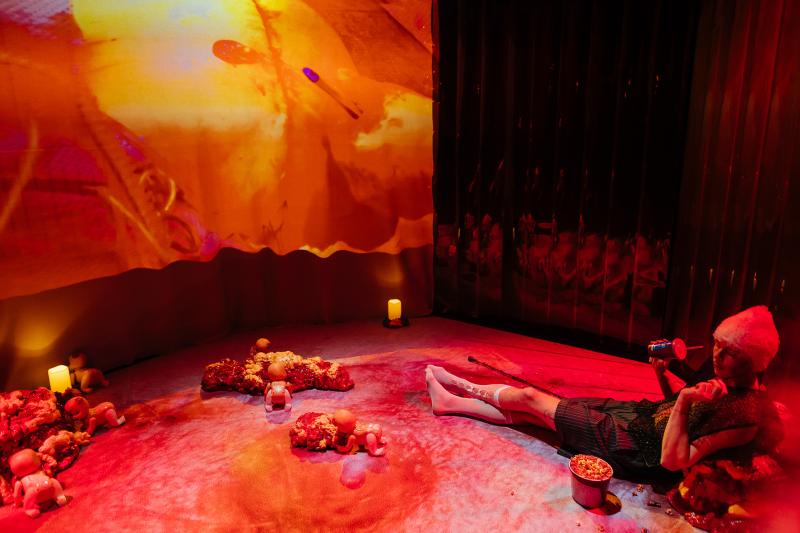 Performer lying on a red-lit stage floor surrounded by toy baby figures and organic sculptural forms, with a large projection behind.