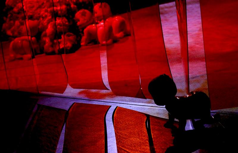 A toy baby figure crawls out from behind a red PVC curtain, with silhouettes of other baby figures and fleshy sculptural objects behind it.