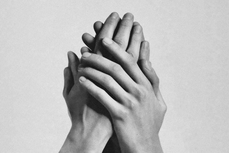Two hands are interlaced and pressed together against a plain light background. The fingers overlap tightly, forming a compact, sculptural shape. The black-and-white photograph emphasizes the contours of the fingers and the soft shadows between them.