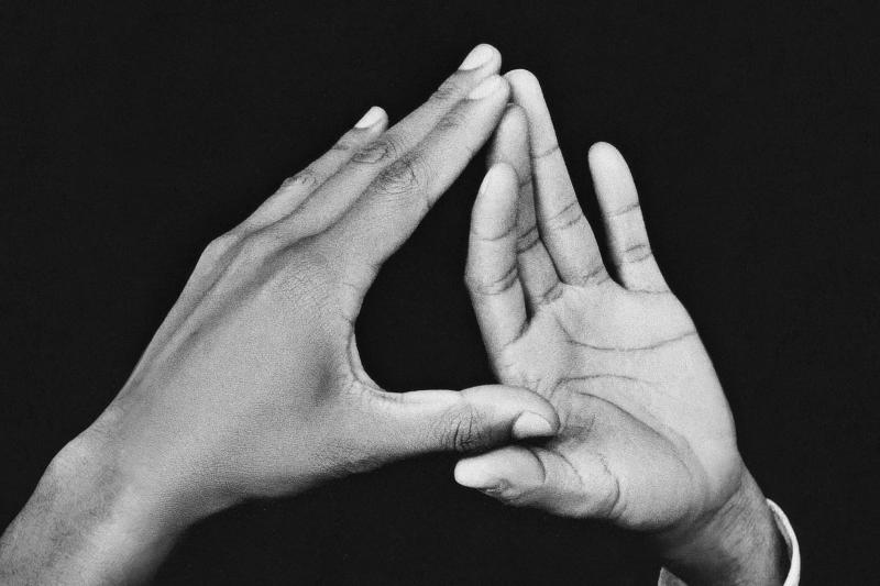 Black-and-white close-up of two human hands against a dark background, positioned so the fingertips touch and form a triangular shape. One hand is shown from the back while the other faces palm-forward, with the thumbs meeting at the bottom and the index fingers meeting at the top.