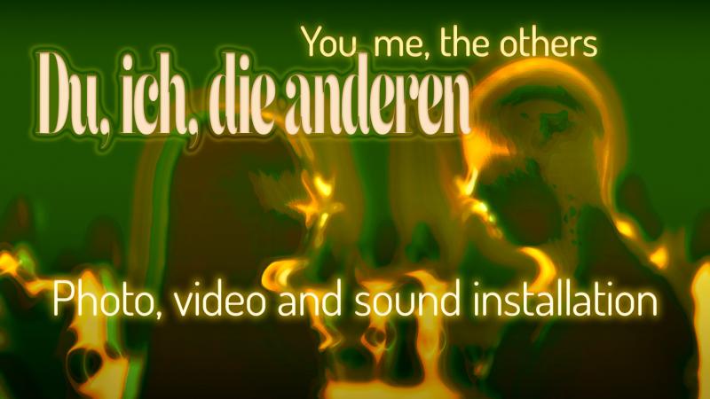 Auf dem Bildd sind zwei verschwommene Personen zu sehen, die von Licht umgeben sind. Im Hintergrund sieht man verschwommene Lichter und Silhouetten. Der Text sagst, du, ich, die anderen. You, me, the others. Photo, video and sound installation.