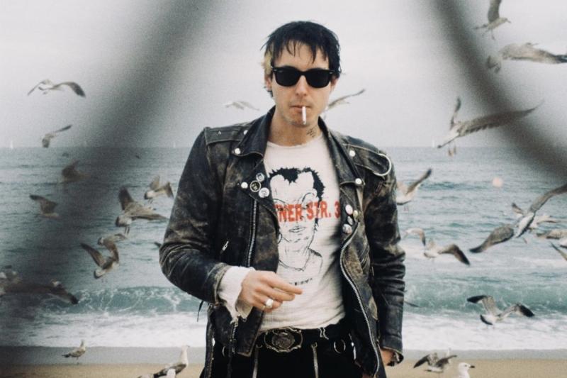 Junger Mann mit Sonnenbrille und nassem Haar raucht Zigarette am Strand. Schwarze Lederjacke, T-Shirt mit rotem Print und Gesichtsmotiv. Viele Möwen im Flug vor grauem Meer. 