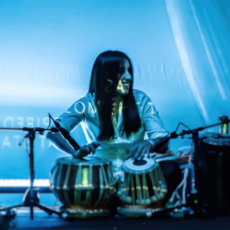 Frau mit langen dunklen Haaren sitzt hinter zwei Tabla-Trommeln und spielt sie vor blauem Hintergrund mit Mikrofonen davor. Woman with long dark hair sitting behind two tabla drums playing them with microphones in front against a blue background