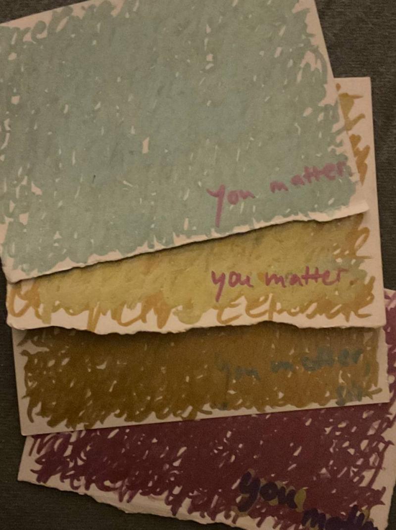 You see four Business Card sized cards laying on top of each other. The first on the top shows green markings with the red sentence „You matter.“ The next card shows golden layered writing with the sentence „You matter.“ The card underneath shows a darker shade of golden layered writing with the sentence „You matter.“ The card at the bottom contains red layering and the sentence „You matter.“ written on it, too.