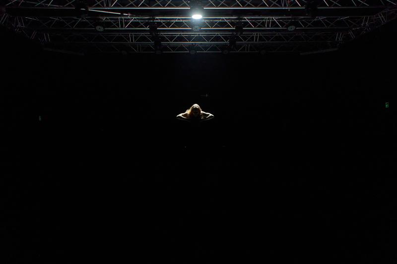 Ein weitestgehend dunkles Bild, ein Spotlight von oben leuchtet auf eine Person, die auf einer dunklen Bühne steht. Nur das Spotlight spendet Licht und zeigt den Kopf, jedoch nicht das Gesicht, sowie die Arme, dessen Ellenbogen zu den Seiten strecken. /// A mainly dark scene: a spotlight from above illuminates a person standing on a dark stage. The spotlight is the only source of light, revealing the head—though not the face—and the arms, with the elbows extending outward to the sides.
