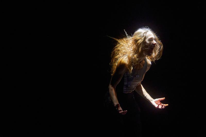 Ein Spotlight von oben leuchtet auf eine Person, die auf einer dunklen Bühne steht. Sie verzieht ihr Gesicht zu einem Schrei, die Haare fliegen, was vermuten lässt, dass sie sich bewegt. Der Körper ist nach rechts ausgerichtet, beide Arme angewinkelt, Ellenbogen an der Hüfte, Unterarme zeigen nach oben, sowie die Handinnenseiten. /// A spotlight from above illuminates a person standing on a dark stage. Her face is contorted in a scream, hair flying, suggesting she is in motion. The body is turned  right. 