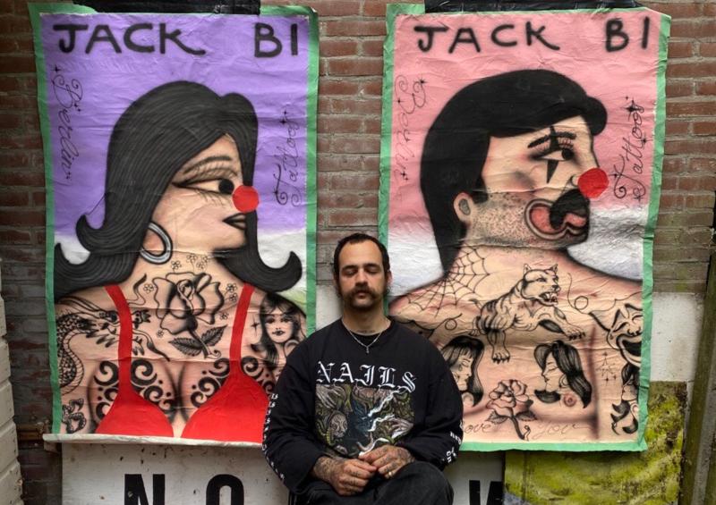 Jack Bi sitzt zwischen zwei seiner Gemälde vor einer Ziegelwand. die Bilder zeigen einen Mann und eine Frau mit Clownsnasen und Tattoos