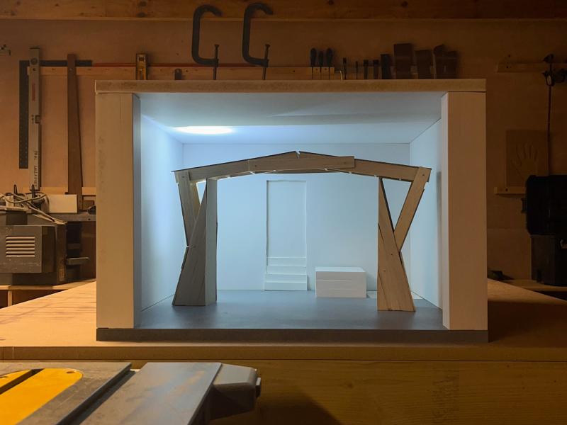 A 1:10 scale model of a gallery sits in a busy workshop. Inside the room stands a monumental gate, lit from behind. The gate perfectly frames a staircase receding into the far wall of the gallery, and a doorway. In the workshop background we see clamps, spirit levels and the emblem of a hand cut into a piece of wood.
