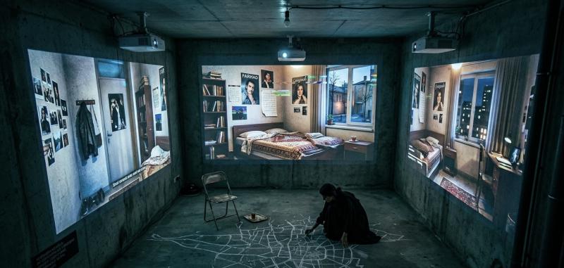 Eine Performerin kniet in einem dunklen Betonraum und zeichnet mit Kreide einen Stadtplan auf den Boden, während an den Wänden fragile Bilder eines Zimmers projiziert werden.