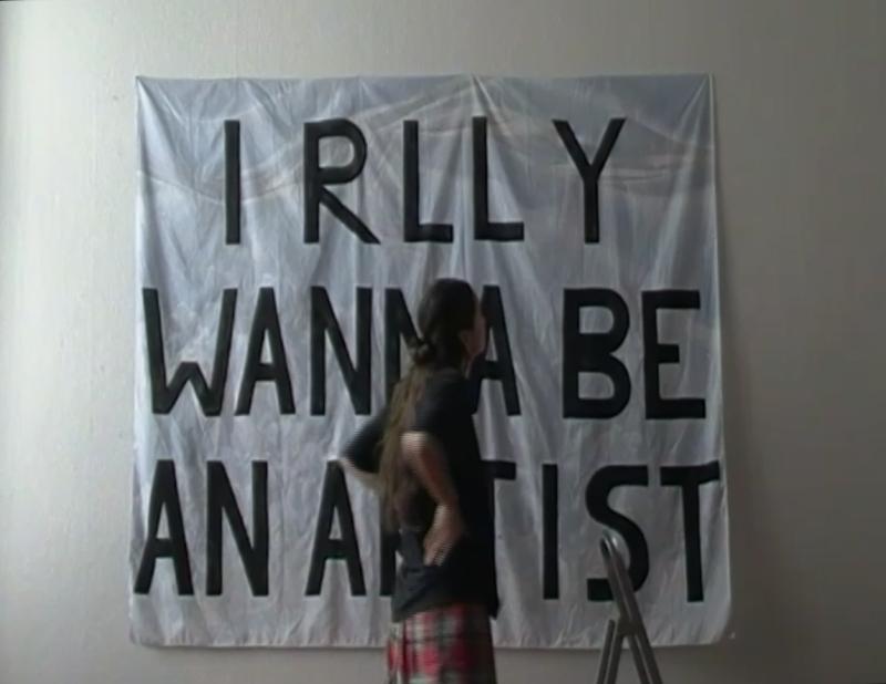 Large white fabric banner with black bold text 'I RLLY WANNA BE AN ARTIST', with woman standing infront.