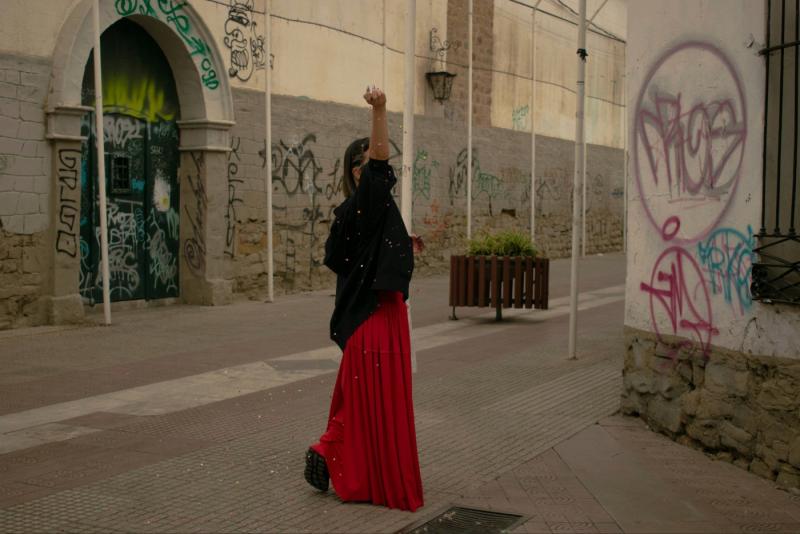 Woman in long red skirt and black hoodie lifting one arm up to throw confetti.