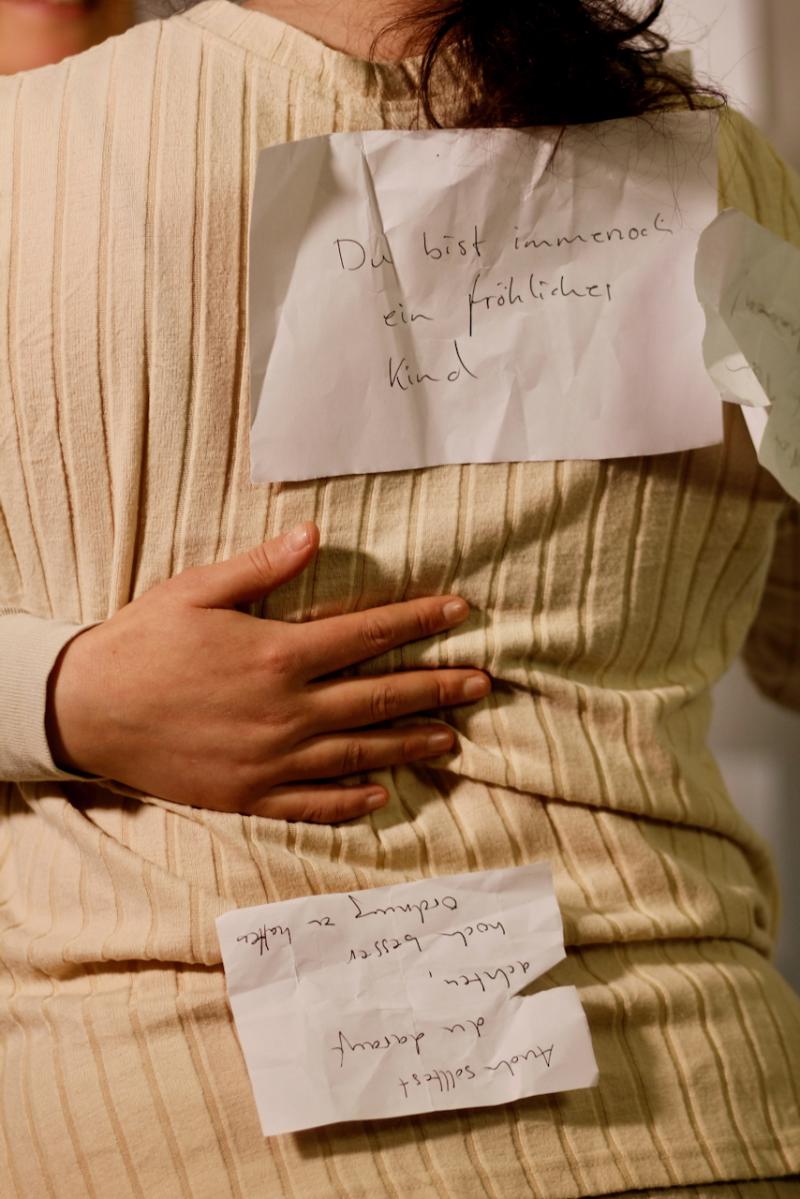 Eine Hand liegt auf dem Rücken der einen Performerinnen, Zettel mit dem Text "Du bist immer noch ein fröhliches Kind" klebt auf dem oberen Rücken