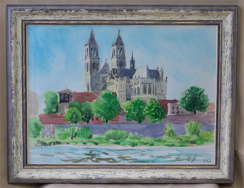 Aquarellgemälde des Magdeburger Doms, der hinter Bäumen und roten Dächern am Ufer eines Flusses steht. Das Bild zeigt eine ruhige Landschaft mit Wasser im Vordergrund und blauem Himmel im Hintergrund und ist in einem hellen, dekorativen Rahmen eingefasst. 