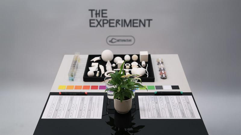 Minimalistisch gestalteter Experiment-Tisch mit geometrischen weißen Formen, Farbkarten, Geruchs– und Geschmacksproben und Fragebögen; in der Mitte steht eine kleine grüne Pflanze. Im Hintergrund der Schriftzug ‚THE EXPERIMENT‘.