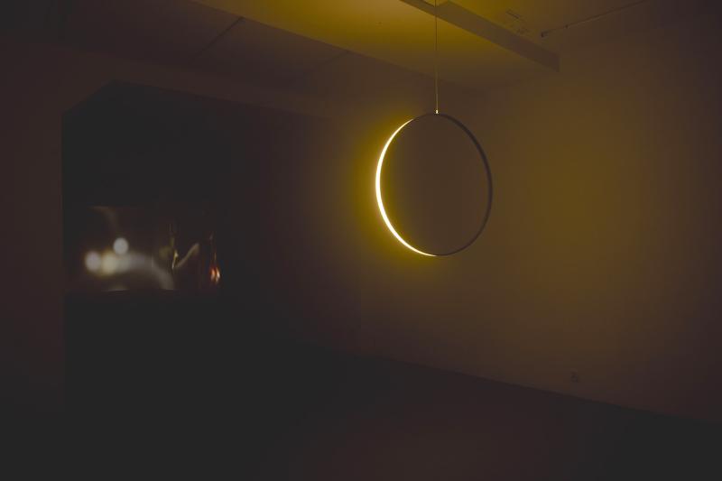A dark exhibition room features a glowing, yellow circular light fixture suspended from the ceiling. In the background, a small video projection is visible on the wall, casting a soft, warm glow throughout the minimalist space.