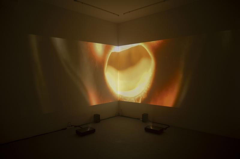 An abstract video projection is displayed across two walls in the corner of a dark room. The image shows a large, glowing, amber-colored circular shape that resembles a close-up of a light source or an organic form, with two projectors visible on the floor below.