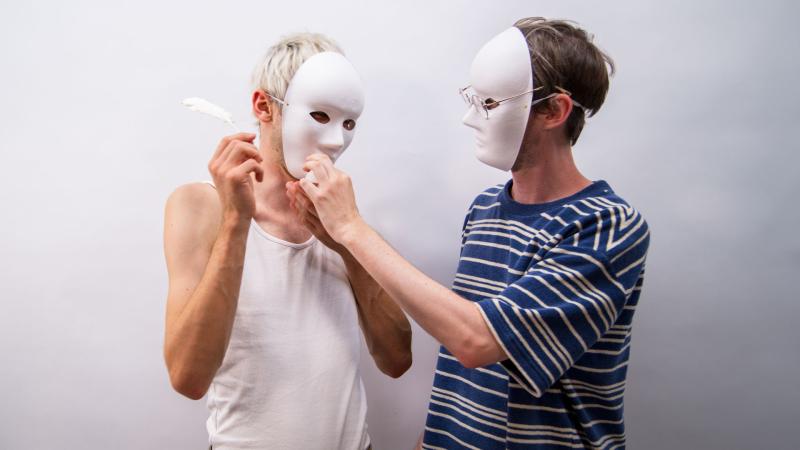 Zwei Personen mit weißen, ausdruckslosen Masken stehen sich einander zugeneigt vor einem weißen Hintergrund gegenüber; eine hält eine Feder, während die andere deren Hand berührt.