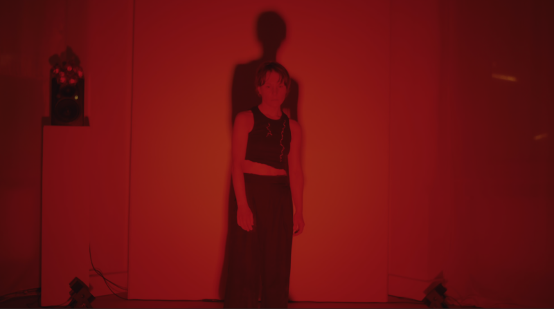 A performer standing in a red light and looking down forward, a shadow forming behind them.