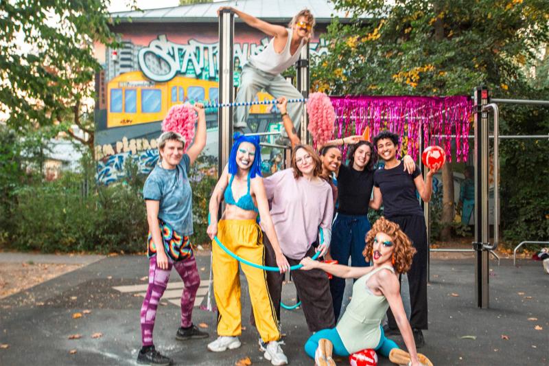Eine Gruppe von sieben bunt gekleideten Menschen posiert fröhlich auf einem Outdoor-Fitnessgerät in einem Park mit Graffiti-Wand im Hintergrund; einige halten Pompons und einen Hula-Hoop-Reifen, während eine Person oben auf der Stange balanciert.