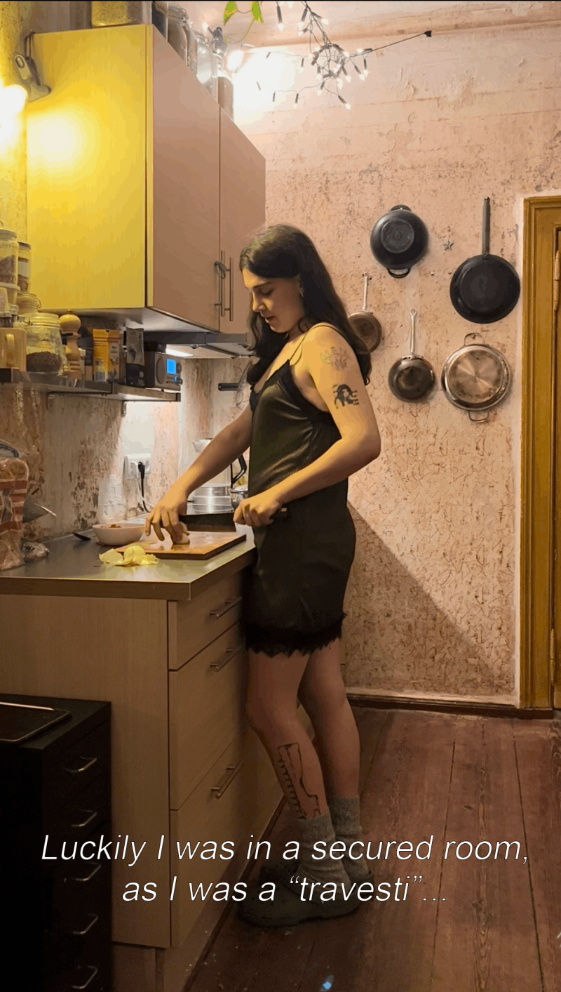 A person in a dark green lace-trim slip dress stands in a kitchen preparing food at the counter. Kitchen utensils hang on a wall in the background. The light is warm, the room feels cozy. Subtitles read: Luckily I was in secured room, as I was a "travesti"...