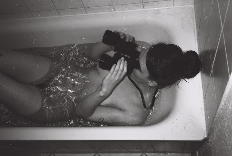 Schwarzweißfotografie einer unbekleideten weiblich-gelesenen Person, die in der Badewanne liegt. Die Person hält ein Fernglas in beiden Händen, durch das sie hindurchschaut. Der Blick ist nach vorne gerichtet. Die Haare sind zu einem Dutt zusammengebunden. Alle Bildteile sind gut erkennbar. Die Badewanne ist mit Wasser gefüllt. Auf Höhe des Bauches zeichnen sich glitzernde Lichtreflexe im Wasser ab und ergeben ein Muster. Das Foto wurde von der Seite aufgenommen und bildet die Person von oben ab.