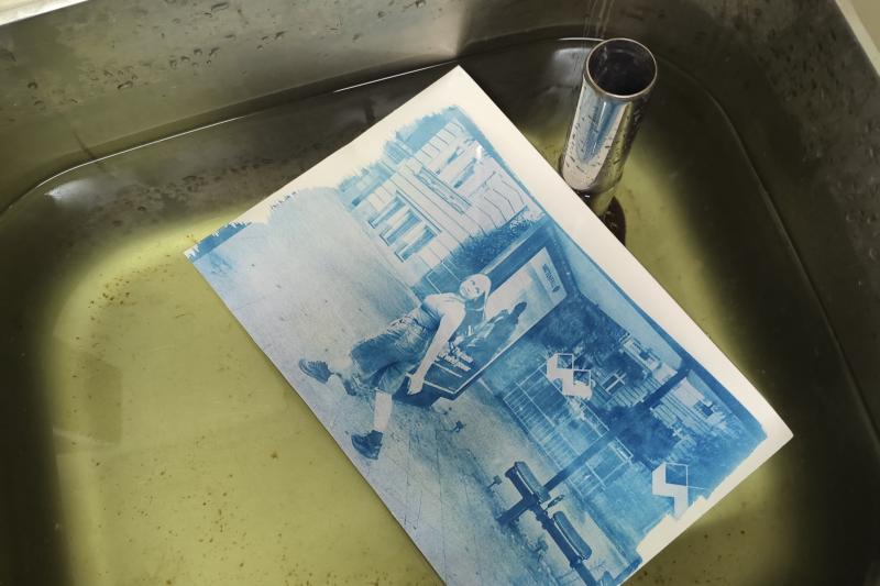 Ein Cyanotypie-Druck schwimmt in einem Wasserbad. Auf dem Abzug ist eine junge Frau abgebildet, die zügig an einer Bushaltestelle vorbeigeht und einen Blick nach hinten wirft.