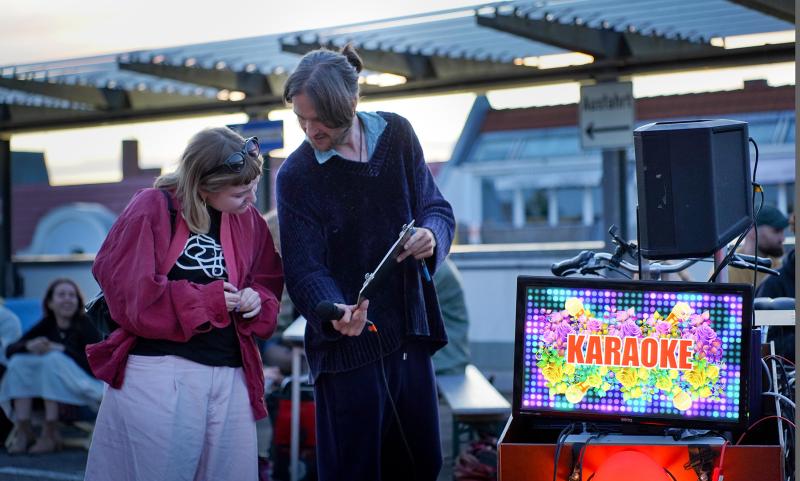 A Karaoke host shares a songlist with a participant by a brightly lit karaoke screen, outdoors.