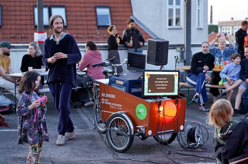 Performer with a cargo bike karaoke setup, including a lyric screen and speakers, surrounded by adults and children at an outdoor community gathering.