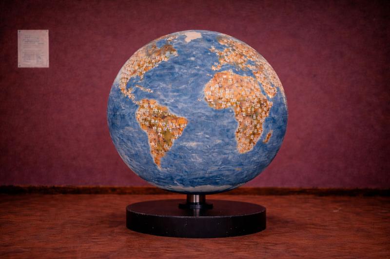 Ein Globus voller multikultureller Porträts bildet eine wachsende menschliche Weltkarte. Jedes Gesicht markiert eine Position zwischen innen, außen und dazwischen — OUT/SIDE/IN — und zeigt, wie Menschen sich bewegen, verbinden und ihren Platz in der Welt neu definieren.