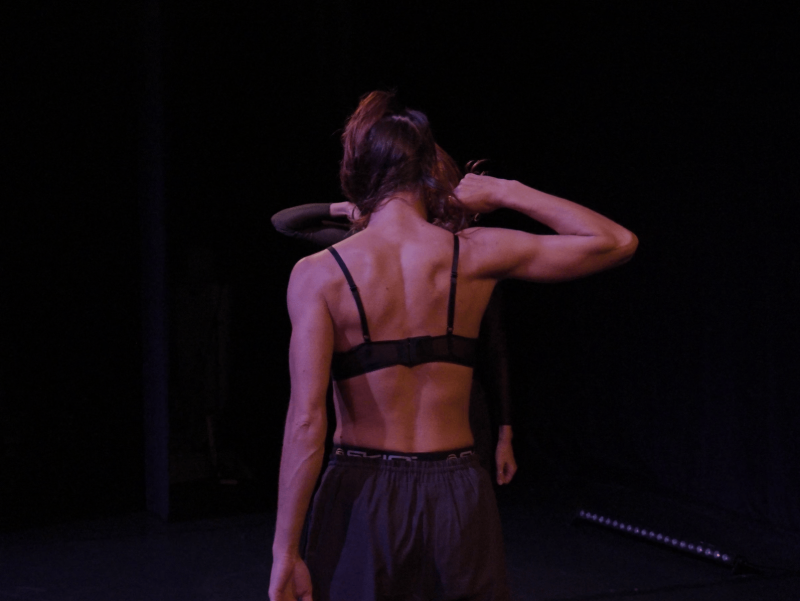A female-read individual stands with her back to the viewer, the left arm hanging down, while the right arm appears to grab the person’s hair, with the elbow pointing to the right. Behind the front person stands another person in the same pose, but facing the first person. Of the person in the back, only the right arm is visible, bent with the elbow pointing to the left; the left arm hangs loosely downward.