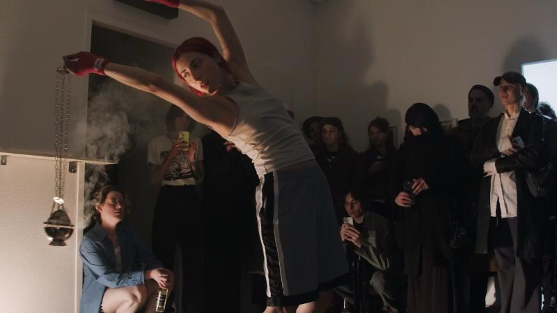 Lisa Kaschubat performing, flexing her body while holding a metal apparatus releasing smoke.