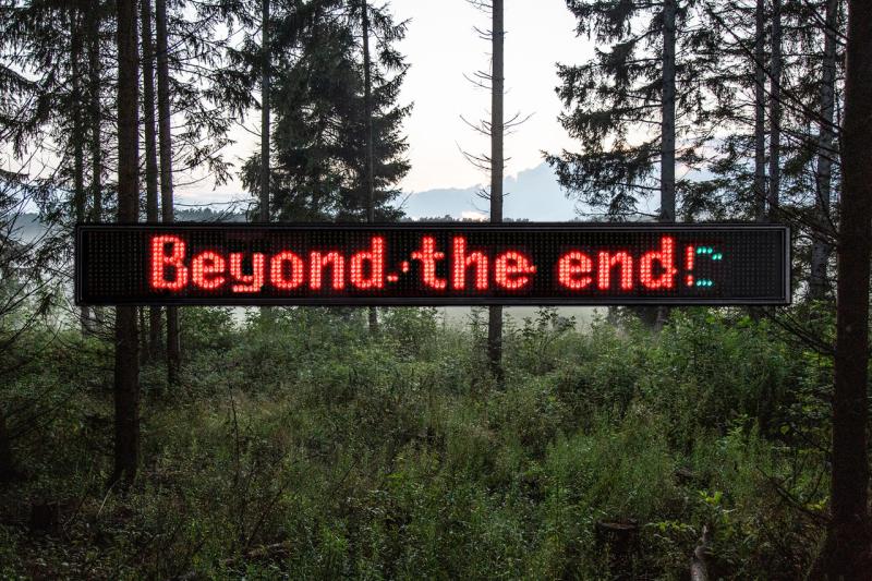 Ein LED-Schild zeigt den Schriftzug "Beyond the end". Es hängt vor einer heimischen Waldlandschaft.