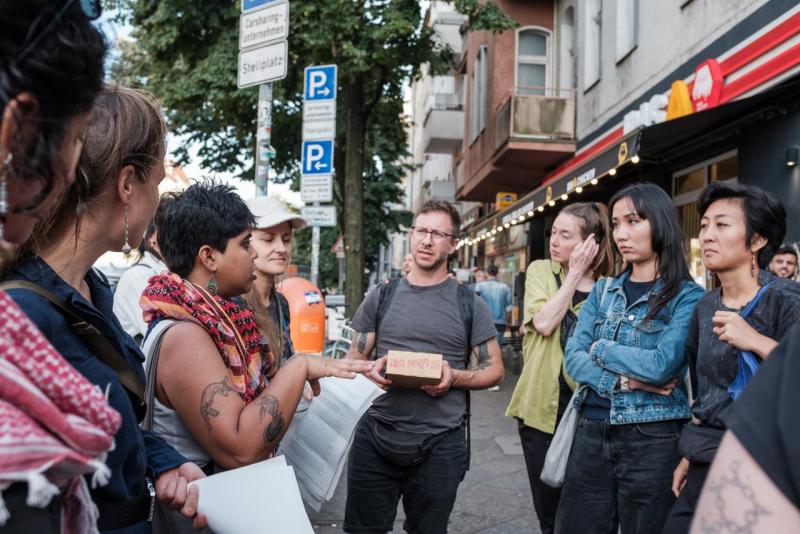 Das Foto zeigt eine Momentaufnahme des Public History Walks auf der Sonnenallee. Die Teilnehmenden stehen in einem Halbkreis und lauschen den beiden Guides.