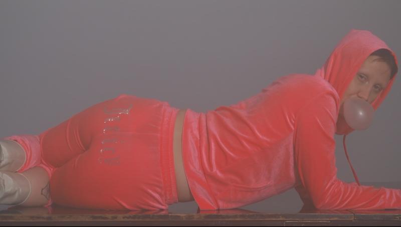 Performerin mit pinkem Jucy Couture Jogging Anzug liegt Kaugummi kauend und mit dem Rücken zur Kamera auf einem Klavier