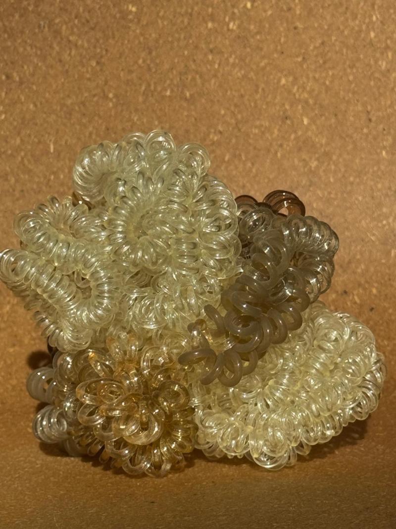 This image shows a cluster of coiled plastic hair ties (spiral hair elastics). The objects are small, spiral-shaped elastic hair bands, often called telephone-cord hair ties because they look like old phone cords. They’re made from translucent plastic and come in slightly different shades (clear, pale yellow, light brown). They’re piled together so the spirals overlap and form flower-like circular shapes.