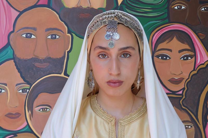 Porträt der Künstlerin Soukaina El Adak mit traditionellem Kopfschmuck und Schmuck, frontal in die Kamera blickend. Im Hintergrund ist ein farbenfrohes Gemälde der Künstlerin mit stilisierten Gesichtern verschiedener Menschen zu sehen.