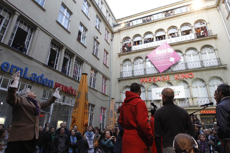 Auf dem Bild stehen viele Menschen in der Passage vor der Neuköllner Oper. Eine Person dirigiert einen Chor, welcher auf dem Balkon der Neuköllner Oper steht und singt. 