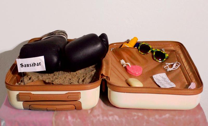 SANSIBAR Frontalansicht; Reisekoffer gefüllt mit Sand und einem Sansibarfläggchen. Dekoriert mit mit Boxhandschuhen, Sonnenbrille, Sonnencreme und Strandsachen.
