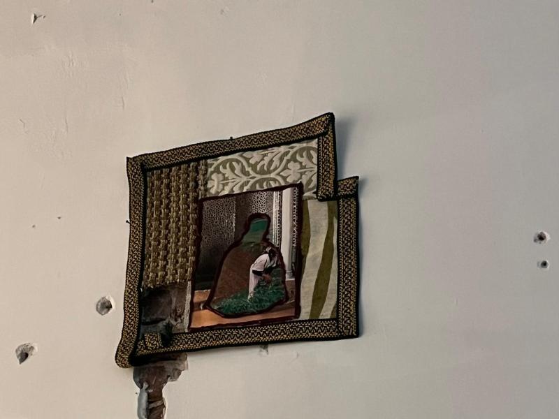 Ein textiles Wandkunstwerk aus zusammengenähten Stoffresten und eingefügten Bildausschnitten hängt an einer weißen Wand. In der Mitte ist eine gerahmte Szene mit einer sitzenden Figur vor ornamentalen Mustern zu sehen. Die Arbeit kombiniert verschiedene Materialien und Strukturen und wirkt wie eine Collage, die kulturelle Darstellungen neu interpretiert.