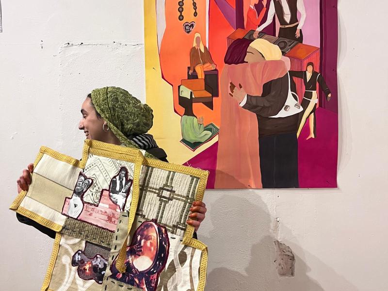 Eine Frau steht seitlich vor einer Wand mit einem farbenfrohen Gemälde und hält ein textiles Kunstwerk mit aufgenähten Bildern und Mustern in den Händen. Sie trägt ein grünes Kopftuch und lächelt, während sie ihre Arbeit präsentiert