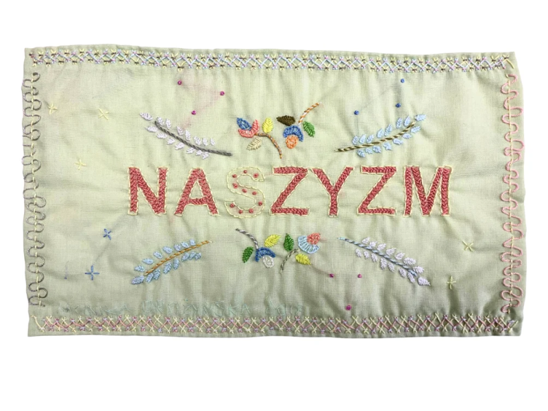 Hellgrüner Stoff mit Stickerei, in der Mitte der rote Schriftzug „NASZYZM“, umgeben von bunten Pflanzenmotiven, Perlen und dekorativen Stichen.